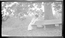 May Gifford sitting on bench under large tree, Oak Lodge Trip
