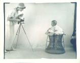 A paper positive showing a woman with a Folding Pocket Kodak camera on a Kodak metal tripod photographing a baby who is on a wicker table