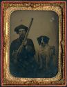 [Seated man with double-barreled shotgun, dog sitting next to him in chair]