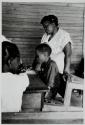 [Children seated at wooden desk, woman (teacher?) standing behind]