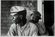 [Boy leaning on man's back, against shingled house]