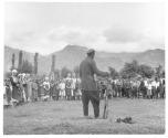 [Man with rifle speaking to semi-circle of children in an open field]
