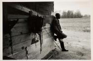 [Man leaning against side of shingled house]