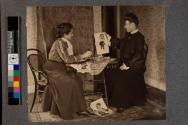 [Two women sitting at table looking at caricatures]