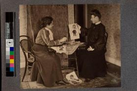 [Two women sitting at table looking at caricatures]