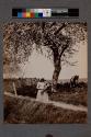 [Girl wearing bonnet holding vegetables with man in shade by tree]