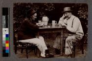 [Two men sitting outside wearing hats and playing poker]