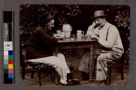 [Two men sitting outside wearing hats and playing poker]