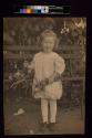 [Young girl holding flowers standing in front of bench]