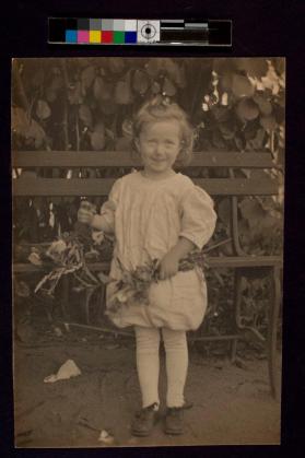 [Young girl holding flowers standing in front of bench]