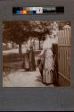 [Two women standing near white fence wearing hats and holding umbrella]