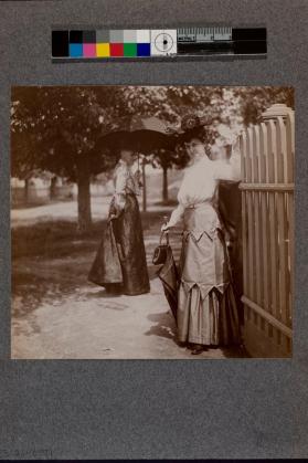 [Two women standing near white fence wearing hats and holding umbrella]