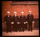 [Four men dressed alike in dark suits and white hats]