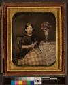 [Girl holding sprig of flowers, vase of flowers on table]