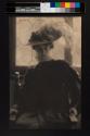 [Olga De Meyer in costume, plumed hat and ruff, by window]