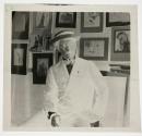 [Man in straw hat seated in front of dispay of framed photos]