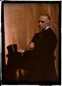 [Man in black, seated in profile holding cigarette, with top hat on knee]