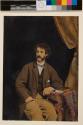 [Young man with curly hair and mutton chops, seated, arm on table with book]