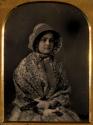 [Seated woman wearing bonnet and print shawl, holding closed fan]