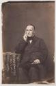 [Late middle-aged man, seated with hand to face, holding a small object]