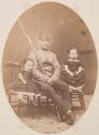 [Soldier, seated on chair  with rifle/bayonet, posed with 2 little girls]