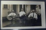 [Three men sitting on lawn in front of house with musical instruments in hand]