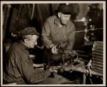 [A gaffer finishing a wide mouthed bottle. The man on the right takes the bottle from the blower on a rod and puts it on a gas flame, which may be seen behind the corrugated tin.]