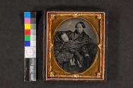[Mother wrapped in shawl with deceased child, man's hand steadies head of large dog at her arm.]