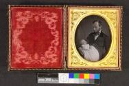 [Man in bowtie holding deceased baby in white]