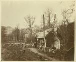 [Man holding an axe near logs in front of a cottage]