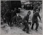 NAZI PRISONERS CARRY WOUNDED COMRADES TO EVACUATION AREA