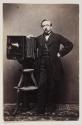 [Man standing beside large portrait camera on stand]
