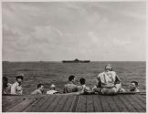 [Military personnel seated by dock watching ships at sea]