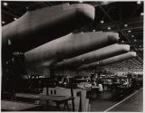 [Interior airplane factory, airplane parts hanging above factory workers]