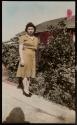 [Woman standing next to bush along sidewalk]