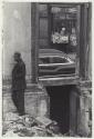 [Man in rubble of building, car seen through doorway]