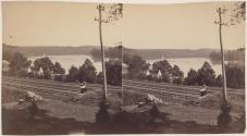 [Man sitting near railroad tracks and telegraph poles near river]