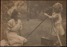 [Woman sitting on ground with child behind camera taking her photograph]