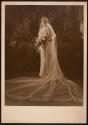 [Standing bride holding bouquet with foliage in background]