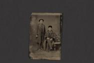 [Two men posing in studio, one seated on bench]