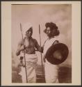 [Large group of mostly children in front of stone wall] \ [Two men holding spears]