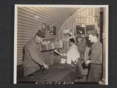 [Tailor and barbershop of a signal company in Northern Ireland. PVT John Tibus, Chicago, ILL., company tailor, pressing the pants of the chief clerk of the signal office T/SGT Donald L. Shristenson, Osseo, Wis. In the background T/5 Adib A. Canamey, Detroit, Mich., gets a haircut before going on a two day pass, by the company barber PVT Zollie Noles, Frolona, GA.  5th Sig Co, 5th Inf Div, Braynsford, County Down, Northren Ireland.]