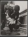[Woman bent over going up stairs with broom, mop, and bucket.]