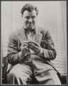 [Man with bow tie seated by window with blinds.]