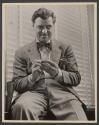[Man with bow tie seated by window with blinds.]