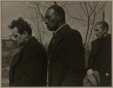[Three men standing in black coats, bare trees and smoke stack in background.]