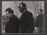 [Three men standing in black coats, bare trees and smoke stack in background.]