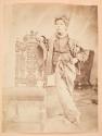 [Young boy standing by ornate carved chair]