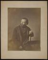 [Bearded man in overcoat, seated in chair]