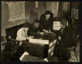 [Woman seated at desk, two men and one woman writing]